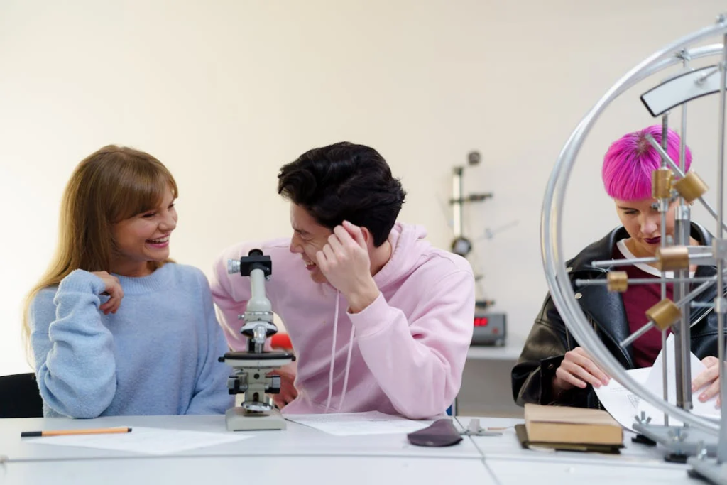 Kündigungs Nachhilfe muster: So gelingt Ihr Weg zur Deutsch-Sprachkompetenz 2 Das Bild zeigt zwei Personen, die an einem Tisch sitzen, wobei eine durch ein Mikroskop schaut. Eine dritte Person mit pinken Haaren ist teilweise rechts im Bild zu sehen, hinter einer Art Fahrradrad-Apparatur. Die Szene deutet auf eine Lernumgebung hin, möglicherweise einen naturwissenschaftlichen Unterricht oder ein Labor, erkennbar am Mikroskop und der experimentellen Einrichtung. Das Bild illustriert eine Bildungsszene, in der Personen wissenschaftliche Beobachtungen oder Experimente durchführen, was auf Lernen, Forschung oder wissenschaftliche Untersuchungen hindeuten könnte.