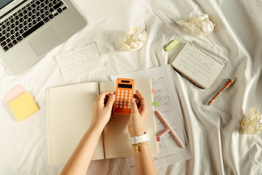 Nachhilfe Mathe Bochum: Verbesserung Ihrer Mathematikkenntnisse in Bochum 2 Studierende löst mathematische Probleme mit einem orangefarbenen Taschenrechner über einem Notizbuch mit handschriftlichen Notizen und Gleichungen, umgeben von zerknülltem Papier, geschlossenem Laptop und weiteren Notizen. Formeln repräsentieren Produktregel und Fundamentalsatz der Analysis.