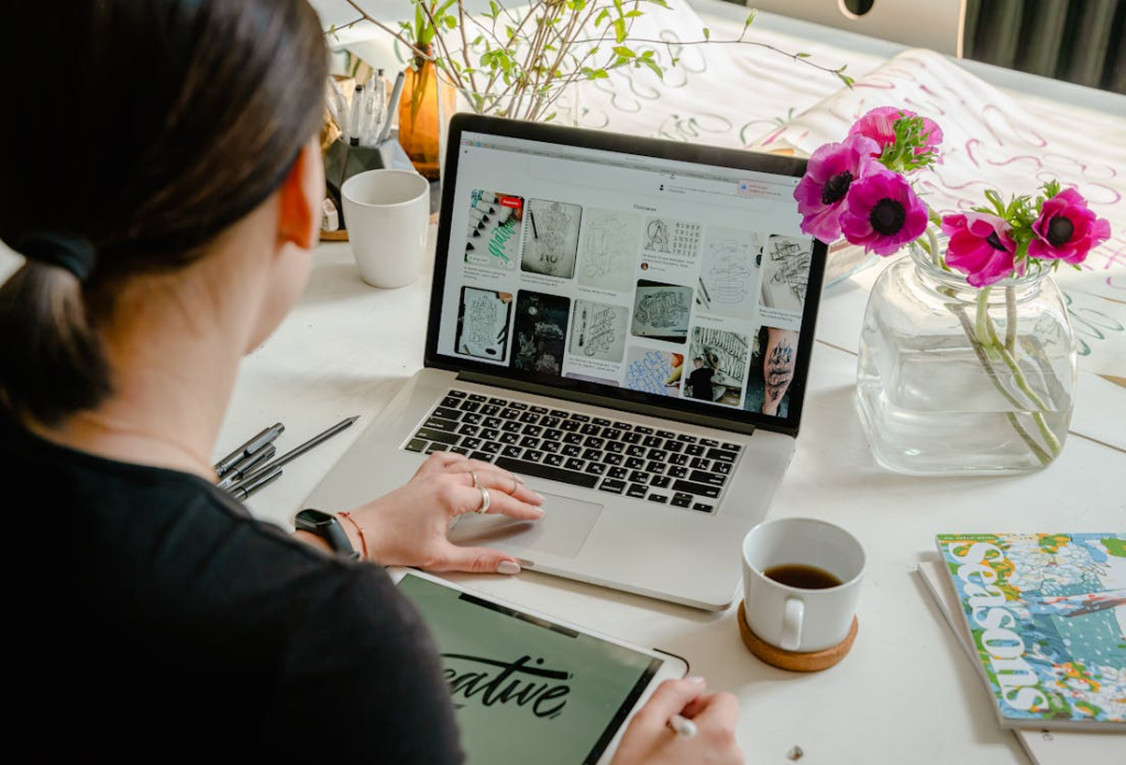Nachhilfe Rechnungswesen meistern: Der ultimative Leitfaden für effektiven Nachhilfeunterricht 2 Das Bild zeigt eine Person von hinten, die an einem Tisch sitzt und an einem Laptop arbeitet, auf dem eine Webseite mit verschiedenen Bildern und Texten zu sehen ist. Neben dem Laptop liegt ein Notizbuch mit handschriftlichen Notizen, ein Stift und ein Smartphone. Vor der Person steht eine klare Glasvase mit rosa Blumen, die ein ästhetisches Element in die Umgebung bringt. Auf dem Tisch ist auch eine Tasse Kaffee oder Tee zu sehen, was auf eine entspannte oder zwanglose Arbeitsumgebung hindeutet. Die Anwesenheit von kreativen Materialien könnte darauf hinweisen, dass die Person in irgendeiner Form von kreativer Arbeit oder Forschung tätig ist.