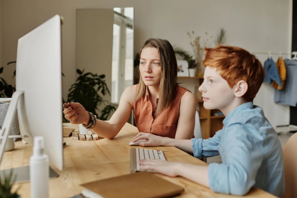 Effektive Mathe-Nachhilfe Leipzig: Ihr Weg zum Erfolg 2 Alt-Text auf Deutsch: Das Bild zeigt zwei Personen, die an einem Schreibtisch sitzen, vor sich einen Desktop-Computer und einige Papiere. Die Person links, ein Erwachsener in einem orangefarbenen Oberteil, hält einen Stift oder Stylus, möglicherweise um mit einem nicht sichtbaren digitalen Gerät zu interagieren. Die Person rechts, vermutlich ein Kind, trägt ein blaues Hemd und blickt auf den Bildschirm des Computers. Die Gesichter beider Personen sind durch braune rechteckige Formen verdeckt. Mathematische oder Hausaufgabenprobleme sind auf diesem Bild nicht zu erkennen.